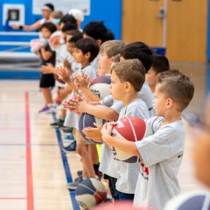 Group of children in matching gray shirts lining up inside a gym to play basketball, eagerly waiting for their turn on the court.